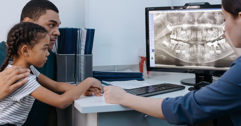 A father and daughter look at a dental X-ray on a screen while a dentist points to the teeth