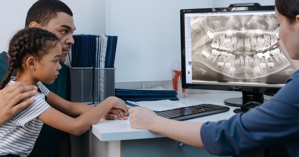 A father and daughter look at a dental X-ray on a screen while a dentist points to the teeth